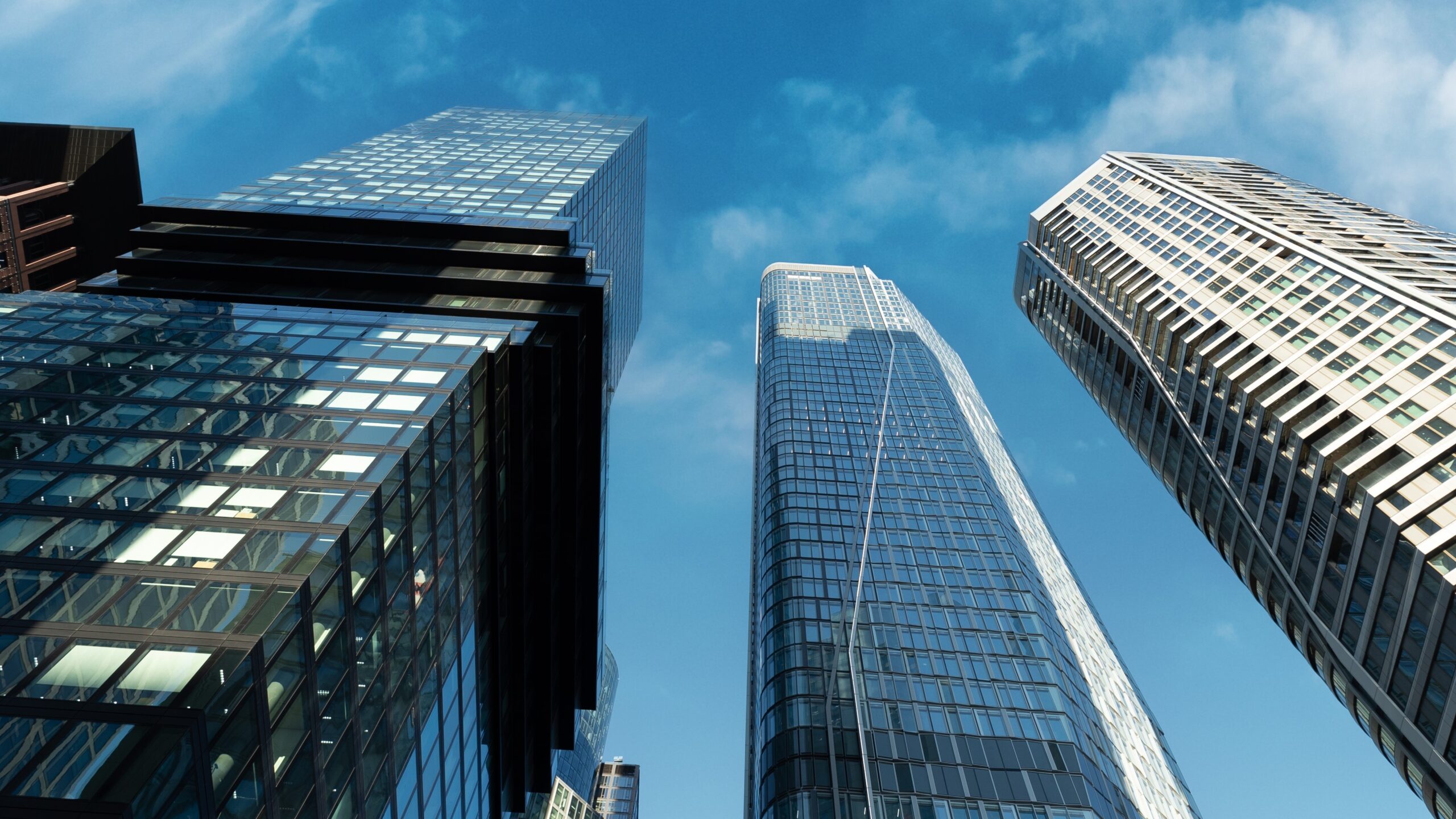 Blick von unten auf moderne Hochhäuser mit Glasfassaden vor einem blauen Himmel mit leichten Wolken. Die Gebäude wirken imposant und repräsentieren eine urbane Architektur.